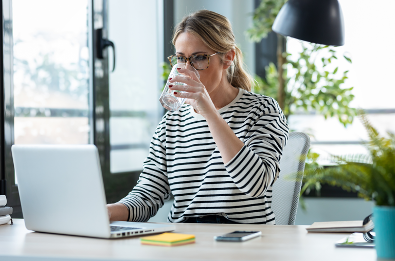 Eine Frau am Schreibtisch vor ihrem Computer trinkt ein Glas Wasser